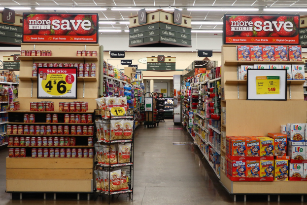 Ein Convenience-Store-Innenraum mit Regalen und Boden voller verschiedener Lebensmittel, beleuchtet von Deckenlampen und mit Informationsschildern mit Text und Zahlen.
