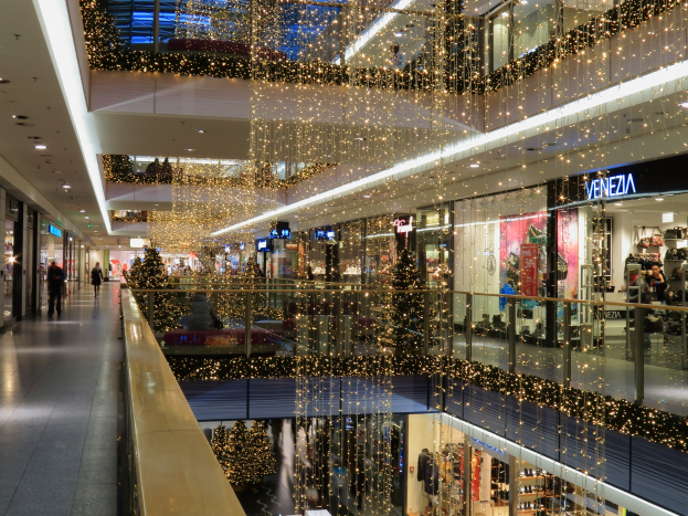 Das Innere eines festlich geschmückten Einkaufszentrums mit Weihnachtsbeleuchtung, das Menschen zeigt, die herumlaufen, Geschäfte mit Schildern, Glasgeländer und von der Decke hängende Lichter.