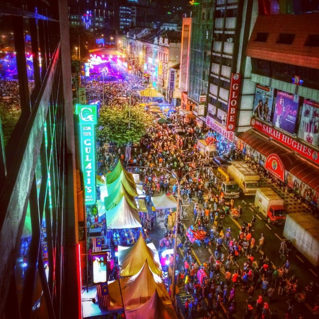 Eine große Menge versammelte sich auf einer Straðe, umgeben von Zelten, Geschäften, Gebäuden und Bannern, mit zahlreichen Lichtern im Hintergrund.