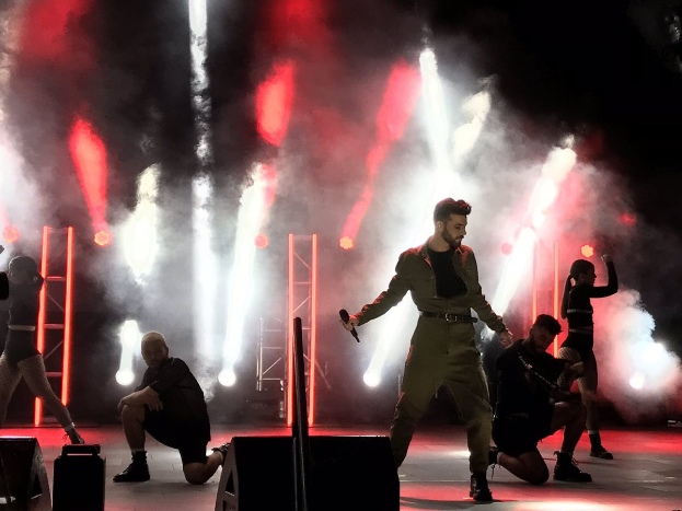 Drake performs on stage at the 2018 Coachella Valley Music and Arts Festival, holding a microphone, surrounded by dancers in a squat position, with speakers, a camera operator, metal poles, and a roof with ceiling lights.