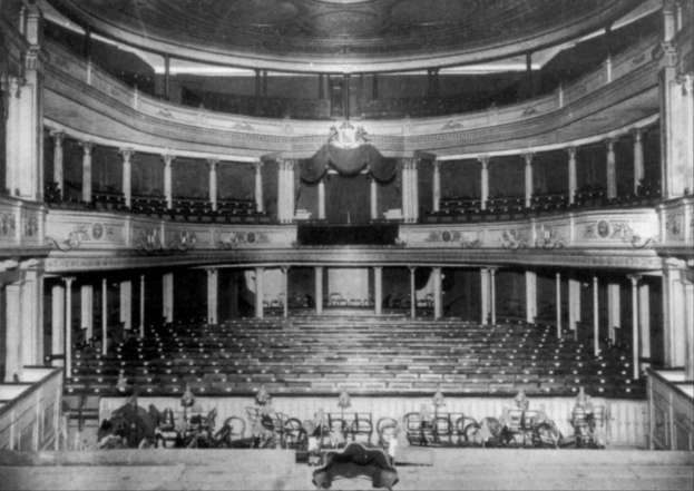 Altes Schwarz-Weiß-Foto des Theatre Royal Auditoriums in London, das Stühle, Treppen, Geländer, Säulen, Lampen und einen Kronleuchter sowie einige sitzende und stehende Personen zeigt.