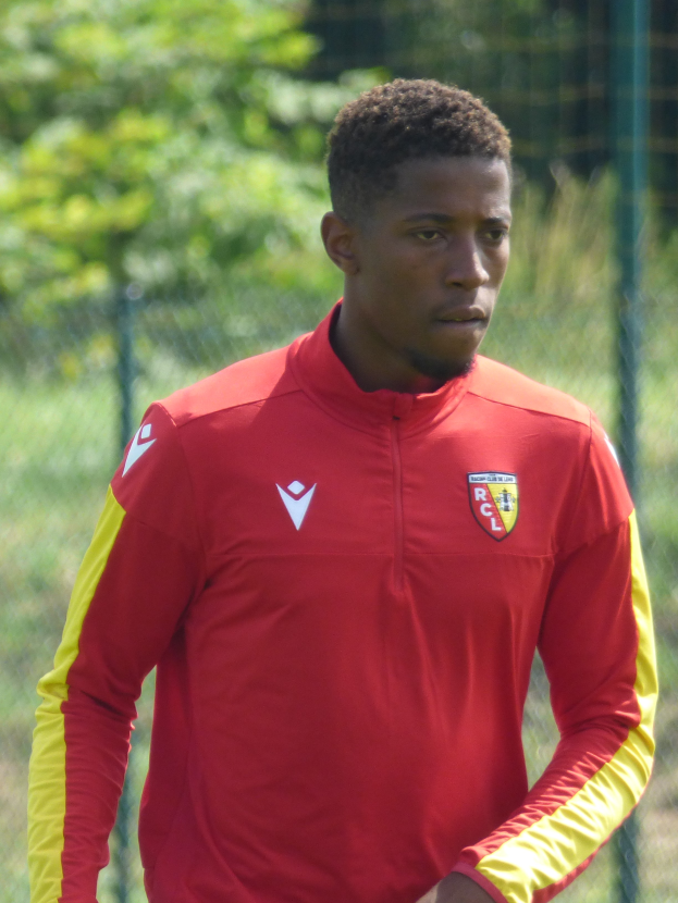 Ein junger Mann in rot-gelbem Trikot steht auf einem Fußballfeld, umgeben von einem Zaun und Bäumen, und sieht entschlossen aus.