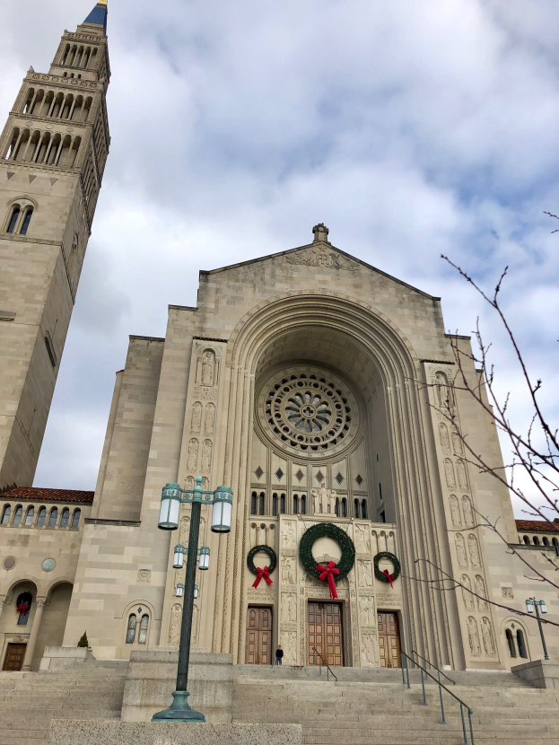 Eine große Kathedrale mit einem zentralen Uhrenturm, geschmückt mit festlichen Kränzen, umgeben von Laternenmasten, Treppen, Geländern und einem Baum auf der rechten Seite, unter einem sichtbaren Himmel.