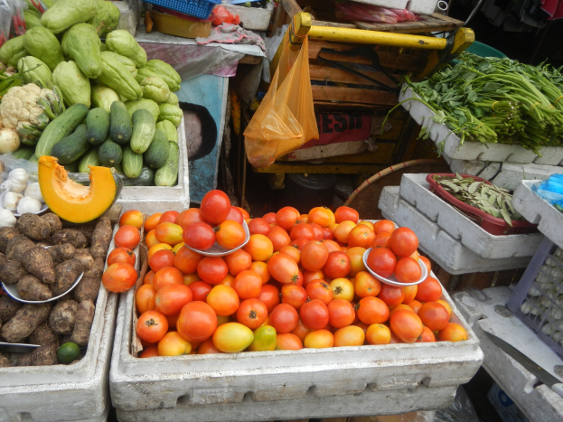 Ein Marktstand mit verschiedenen Gemüsen, darunter Tomaten, Paprika und Blumenkohl, in Schalen mit Plastikabdeckungen und anderen Gegenständen im Hintergrund.