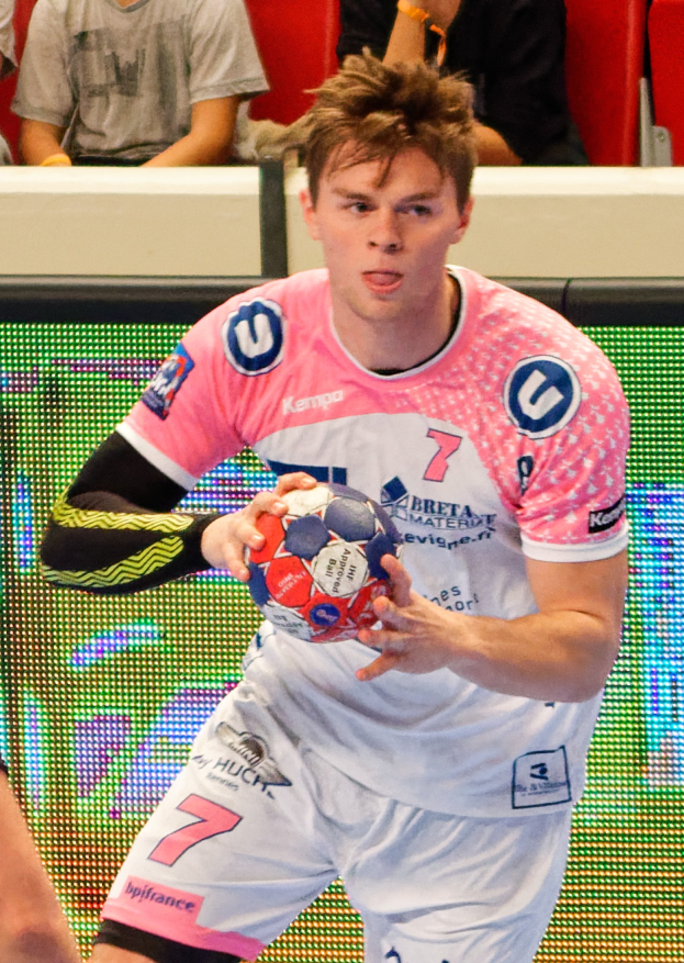 Ein Mann in einem pinken Hemd mit einem Fußball, daneben ist eine andere Person Hand zu sehen und im Hintergrund sitzen Menschen auf Stühlen.