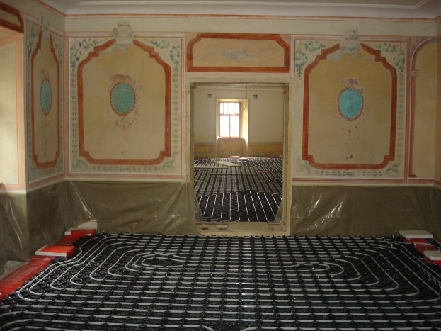 Ein Raum mit einer Fußbodenheizung in der Mitte, umgeben von Wänden und einem Fenster im Hintergrund, mit verschiedenen Gegenständen auf dem Boden, die wahrscheinlich mit dem Heizungssystem zusammenhängen.