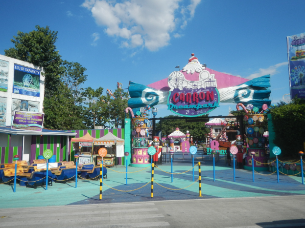 Karussell am Eingang eines Parks, mit Straße, Umzäunung, Buden, Banner, Bäumen und einem bewölkten Himmel im Hintergrund.