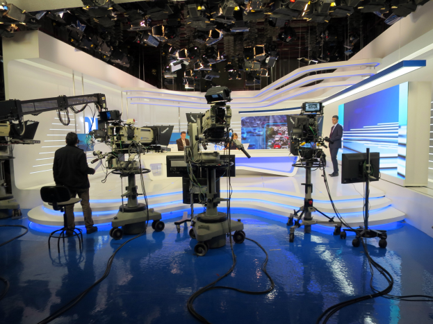 Ein Fernsehstudio mit mehreren Kameras auf dem Boden, einer sitzenden Person im Vordergrund und einigen stehenden Individuen im Hintergrund, mit decke montierten Lampen für eine Livesendung.