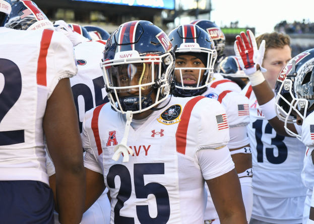 Gruppe von Männern in Football-Outfits auf einem Feld mit Helmen und Handschuhen, mit einer unscharfen Menge, einem Anzeigebrett mit 'Auburn vs Virginia Tech - 11/28/15 College Football Pick, Odds, and Prediction' und Himmel im Hintergrund.