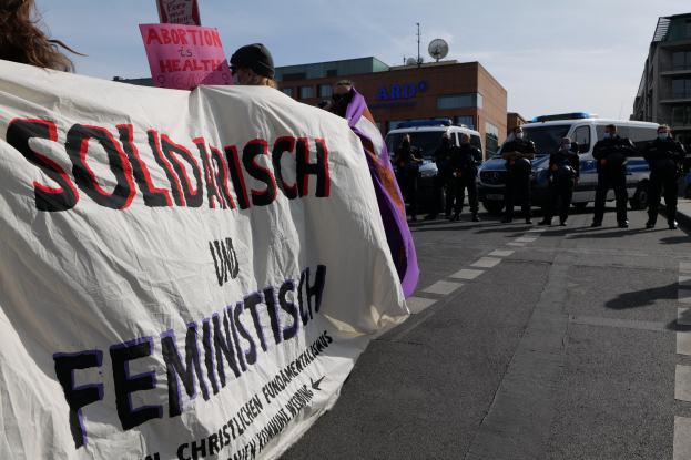 Gruppe von Menschen marschiert auf einer Straße und hält eine 'Solidarität und Feminismus'-Schleife, mit parkenden Fahrzeugen und Gebäuden im Hintergrund unter einem klaren blauen Himmel.