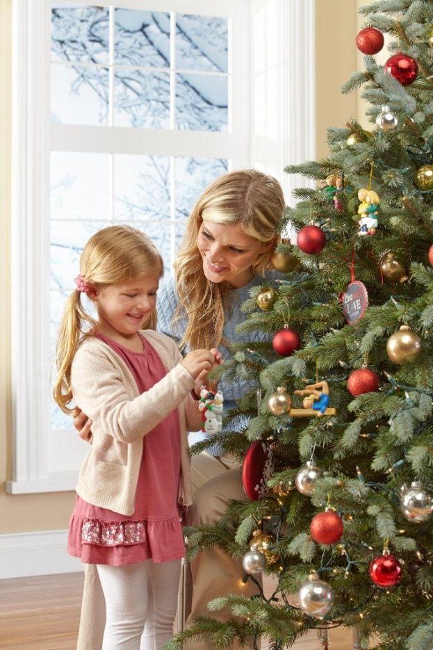 Ein geschm├╝ckter Weihnachtsbaum steht im Vordergrund eines Raums, mit einer Frau und einem Kind in der N├Ąhe; ein Fenster und eine Wand sind im Hintergrund zu sehen, mit Tannenb├Ąumen im Freien.