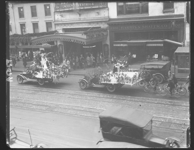 Ein Schwarz-Weiß-Foto eines Umzugs auf einer Stadtstraße mit Fahrzeugen, Fußgängern und Gebäuden im Hintergrund.