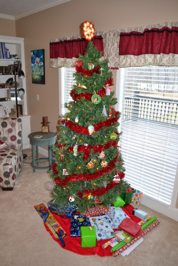 Ein Weihnachtsbaum mit Geschenken vorne, eine Couch, ein Tisch, ein Bücherregal, ein wandhängendes Bild und eine Stehlampe mit Stange, alles in einem Raum mit Fenstern und Vorhängen.