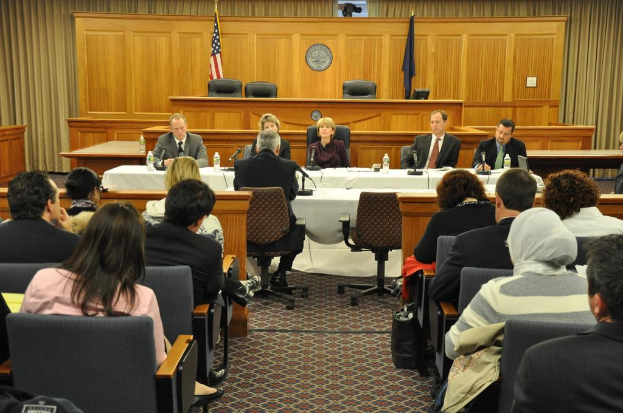 Ein Versammlungsraum mit einer Gruppe von Menschen, die auf Stühlen sitzen, zwei Flaggen anderer Länder, ein orangefarbener Schreibtisch mit Wasserflaschen und einem Mikrofon.