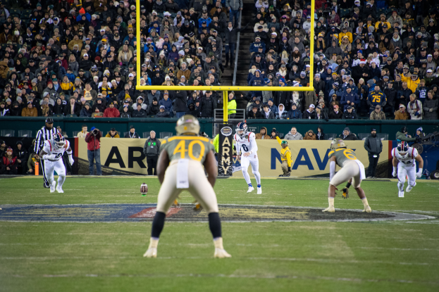 Ein Fussballspiel wird in einem Stadion mit Spielern in Sportkleidung und Helmen im Vordergrund und einer Menge von Zuschauern im Hintergrund sowie sichtbaren Pfählen und Tafeln mit Text gespielt.