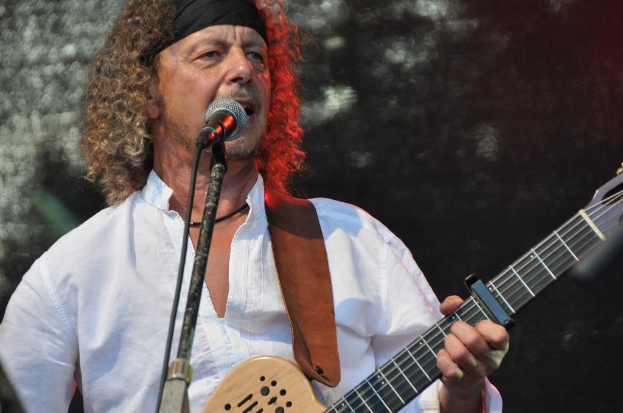 Mann in weißem Hemd und schwarzem Stirnband singt in ein Mikrofon und hält eine Gitarre.