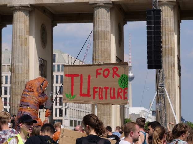 Eine Gruppe von Menschen steht vor dem Brandenburger Tor in Berlin, Deutschland, wobei eine Person in Kostüm mit einem Schild steht, Bäume und Gebäude im Hintergrund unter einem bewölkten Himmel.