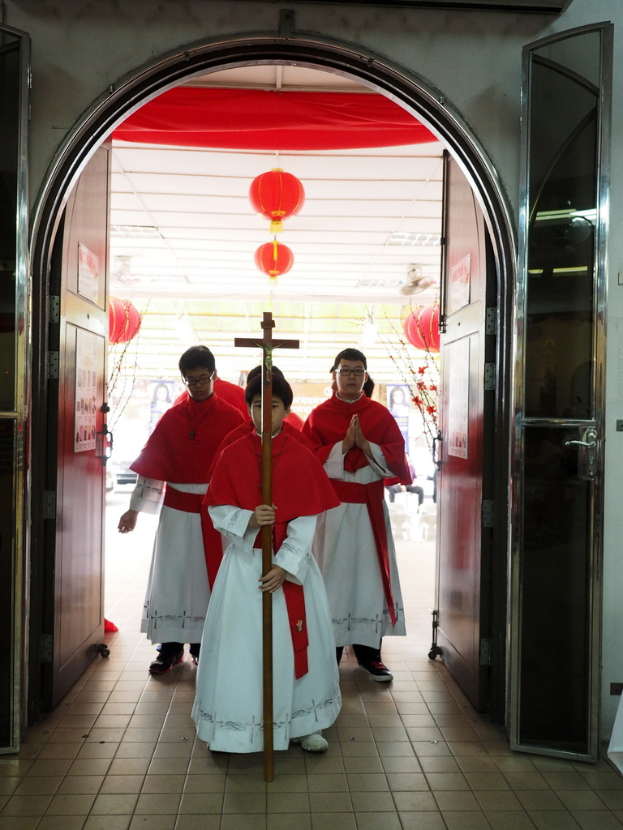 Menschen gehen mit einem Kreuz in einer Kathedrale, mit Türen links und rechts und Blumen und dekorativen Elementen im Hintergrund.