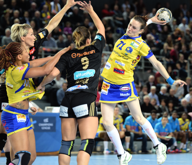 Eine Gruppe Frauen beim Handballspielen, eine hält den Ball, mit unscharfen Zuschauern und Text im Hintergrund.