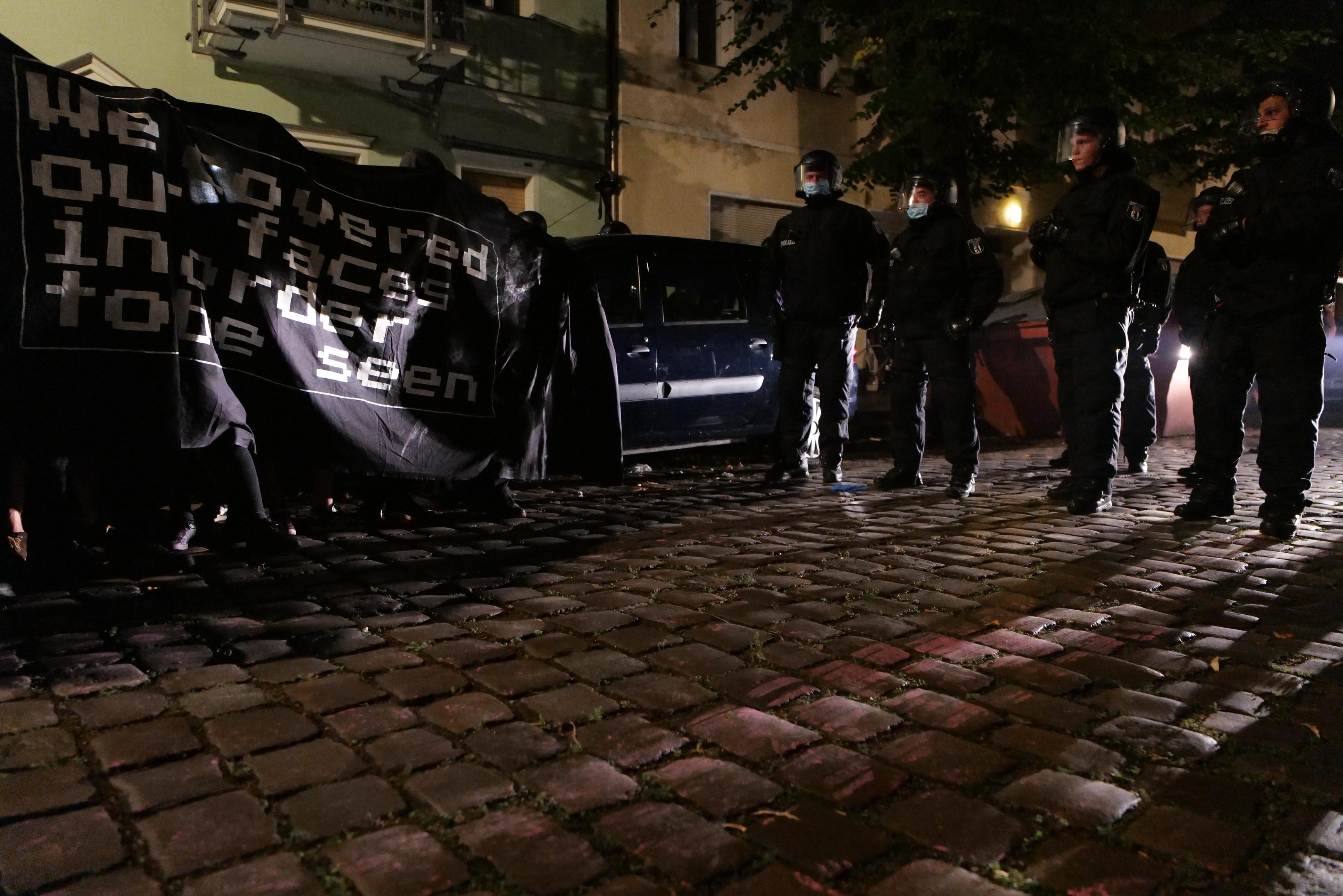 Eine Gruppe von Polizisten in Helmen steht auf einer Kopfsteinpflasterstraße in der Nacht, hält ein Banner, mit Fahrzeugen, Gebäuden, Bäumen und Straßenbeleuchtung im Hintergrund.