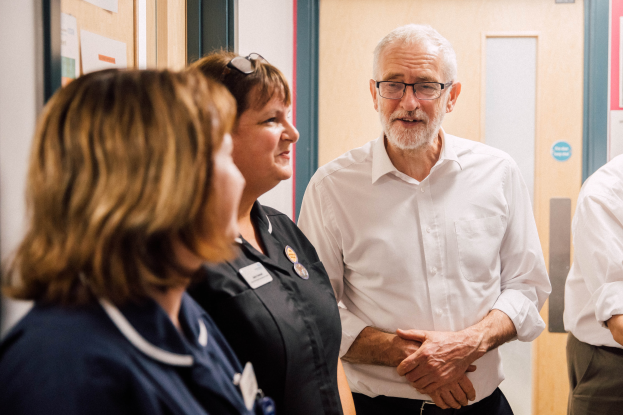 Eine Gruppe von Menschen, die in einem Raum mit einer Tür im Hintergrund und einem Anschlagbrett mit Papieren an der linken Seite steht und spricht.
