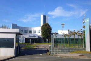 Ein großes, mehrstöckiges Gebäude mit zahlreichen Fenstern und einem Tor an der Vorderseite, umgeben von Straßenmasten, Straßenlaternen, Gittern, Bäumen, Gras und einem bewölkten Himmel, das als Hauptsitz von Iberostica in den Niederlanden dient.