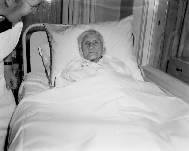 Ein älteres Weib liegt in einem Krankenhausbett mit einem weißen Laken und Kissen, während eine Krankenschwester neben ihr steht, mit einem Fenster und Vorhängen im Hintergrund.