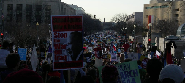Menschen auf einer Straße mit Plakaten, mit Gebäuden, Bäumen, Laternen und dem Himmel im Hintergrund.