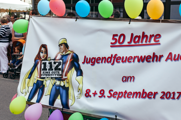 Eine Gruppe von Menschen auf einer Straße, einige mit Kinderwagen, mit einem Banner mit der Aufschrift "50 Jahre Jugendfeuerwehr August 2017" mit Text- und Cartoons, einem Zaun, Ballons, einem Zelt, einem Gebäude mit Fenstern und Bäumen im Hintergrund.