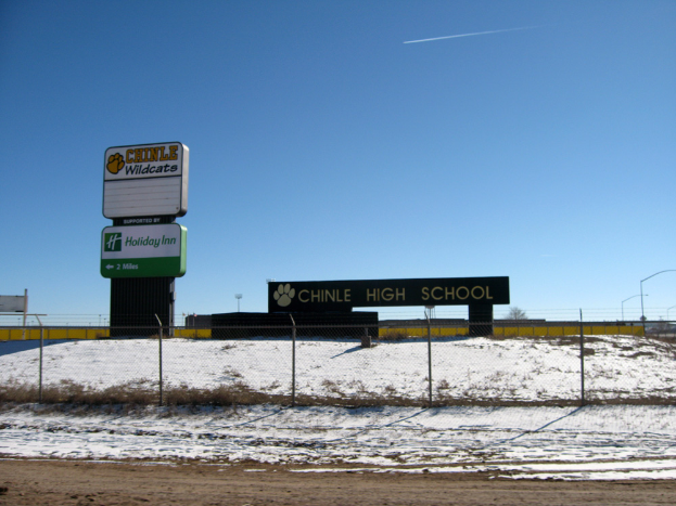 Eine schneebedeckte Straße mit einem Zaun, der Text anzeigt, einer Wand, Laternenpfählen, Bäumen und einem klaren Himmel, mit einem Schild, das "Chinle High School" im Vordergrund anzeigt.