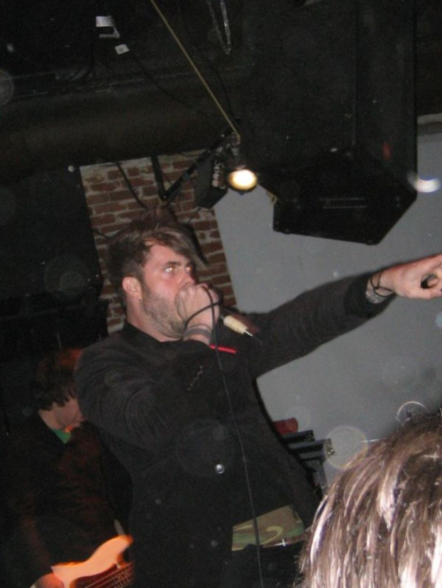 Ein Mann singt in ein Mikrofon in der Mitte einer Gruppe von Menschen, mit sichtbaren Lichtern im Hintergrund.