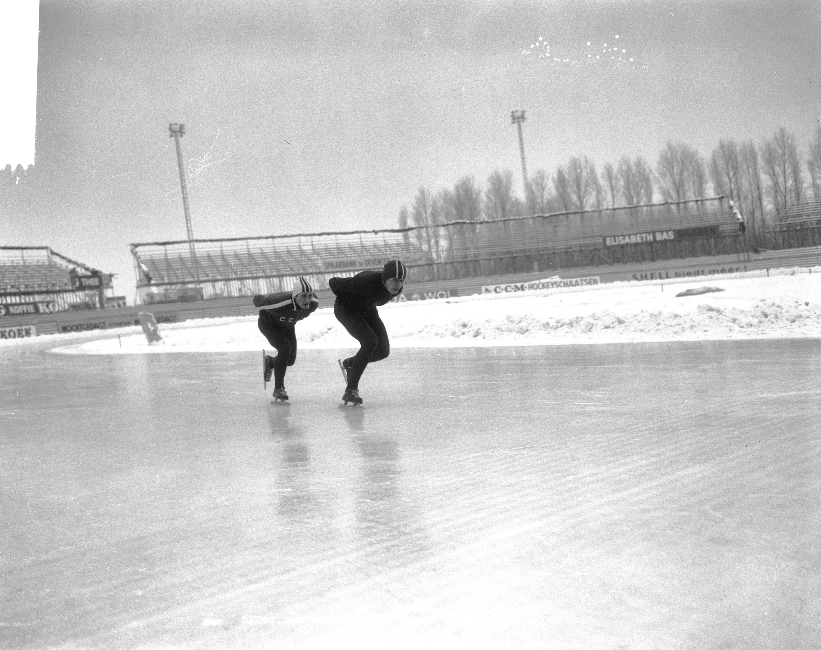 Zwei Menschen, die auf einer Eisbahn im Schnee Schlittschuh laufen, tragen Helme und Schlittschuhe, im Hintergrund sind Bäume, Pfosten, ein Zaun und der Himmel zu sehen.