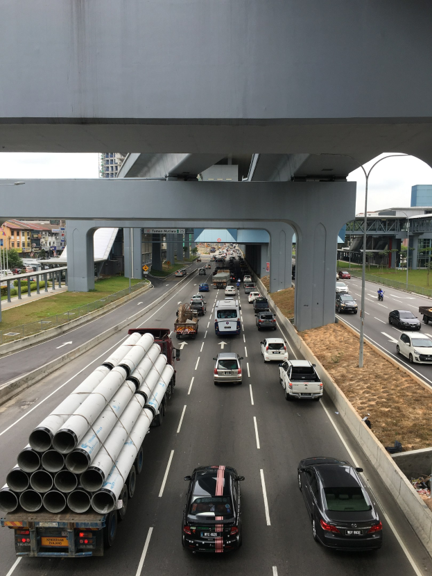 Eine vielbefahrene Autobahn mit mehreren Fahrzeugen, einer Brücke darüber, Straßenlaternen, Gras, Gebäuden, Bäumen und dem Himmel.