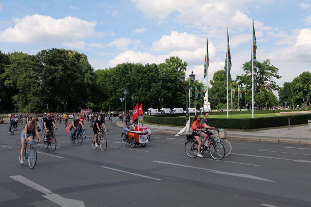 Gruppe von Menschen, die auf Fahrrädern eine von Bäumen gesäumte Straße mit Laternenmasten und Fahnenmasten entlangfahren, während der Pariser Fahrradtour 2016, mit bewölktem Himmel und Fahrzeugen im Hintergrund.