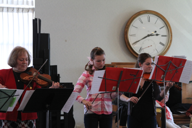 Drei Frauen auf der Bühne mit Geigen stehend und Musiknotenständer vor sich, während eine Gruppe von sitzenden Frauen im Vordergrund zuhört; eine Uhr hängt an der weißen Wand im Hintergrund, mit einem weißen Vorhang in der linken Ecke.