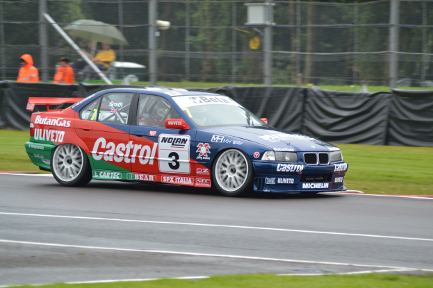 Ein Auto fährt auf einer Rennstrecke mit Gras auf beiden Seiten und einem Maschendrahtzaun, der mit schwarzem Stoff bedeckt ist, und Menschen in orangefarbenen Jacken mit Schirmen im Hintergrund.