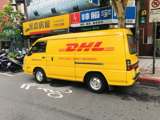 Gelber DHL-Lieferwagen auf der Straße geparkt, umgeben von Gebäuden, Bäumen, Schildern, Strommasten, Ampeln und einem Fußgänger auf dem Bürgersteig.