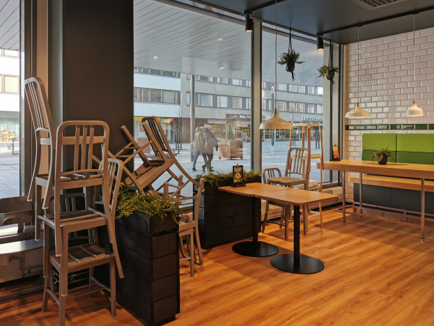 Ein Restaurant mit hölzernen Tischen und Stühlen vor einem großen Fenster, umgeben von Pflanzen und Lichtern, mit Menschen und Gebäuden sichtbar außerhalb in Berlin, Deutschland.