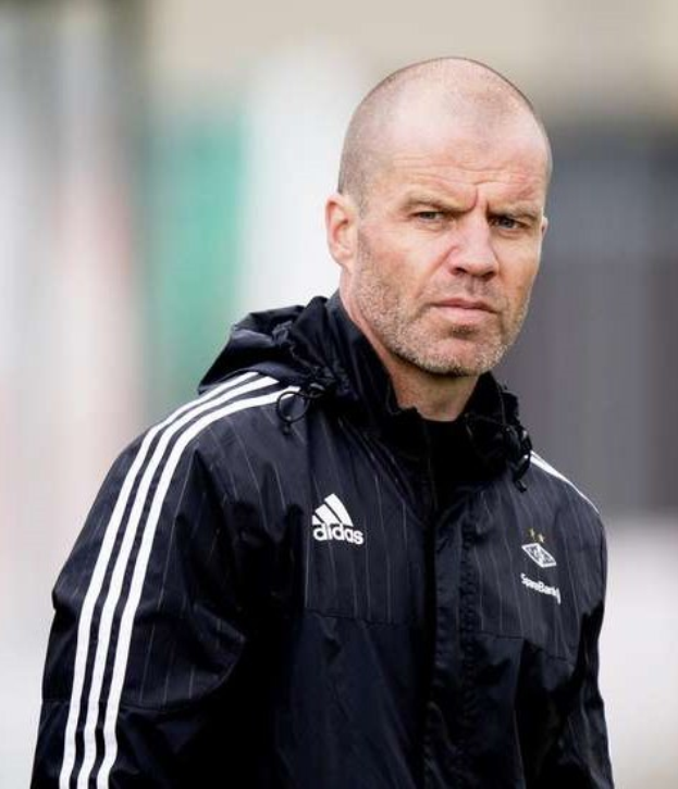 Ein kahlk├Âpfiger Mann in einer schwarzen Jacke steht auf einem Fuballfeld, mit einer marineblauen Jacke mit wei├čen Streifen und einem wei├čen Kragen, die H├Ąnde verschr├Ąnkt und einen ernsten Ausdruck.