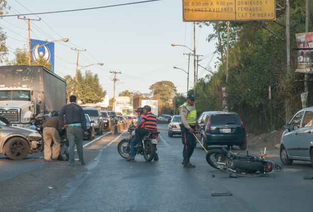 Eine Gruppe von Menschen um ein verunglücktes Motorrad auf der Straße herumstehend mit mehreren Fahrzeugen, darunter ein Lastwagen, und einem Hintergrund aus Bäumen, Pfählen, Lampen und Schildern unter einem Himmel.