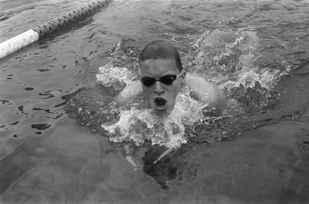 Ein Mann schwimmt in einem Pool mit offenem Mund und trägt eine Schwimmbrille, umgeben von Bahntrennern, auf einem Schwarz-Weiß-Bild.