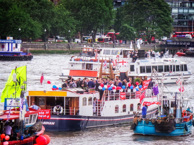Eine Gruppe von Booten mit Menschen, Flaggen und Luftballons auf einem Gewässer, mit Bäumen, Fahrzeugen, Laternenmasten und Gebäuden im Hintergrund.