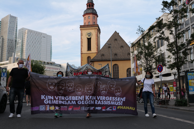 Eine Gruppe maskierter Menschen marschiert mit Protestschildern die Straße entlang, mit einem geparkten Auto auf der linken Seite, Gebäuden, Bäumen, Schildern, Pfosten und einem Uhrenturm im Hintergrund unter einem klaren blauen Himmel.