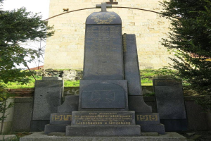 Ein Denkmal für deutsche Soldaten, die im Zweiten Weltkrieg starben, vor einer Kirche mit einem Kreuz, umgeben von Bäumen und einer Mauer im Hintergrund.
