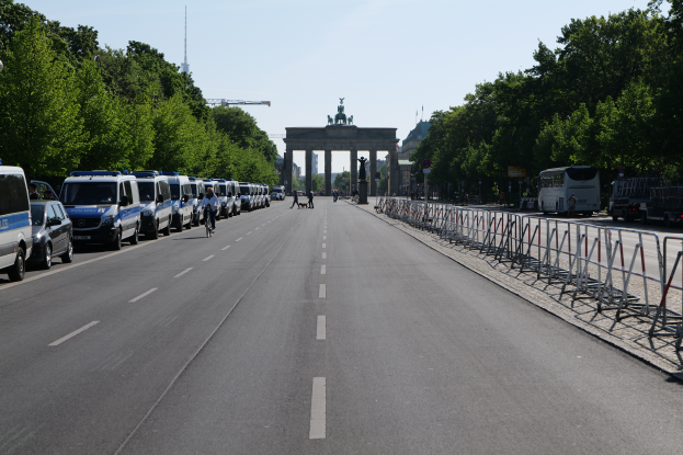 Eine Reihe von Polizeiwagen, die auf einer Straße vor dem Brandenburger Tor in Berlin, Deutschland, geparkt sind, mit Menschen auf Fahrrädern und auf der Straße, Barrieren, Bäumen und einem Bogen mit Statuen im Hintergrund.
