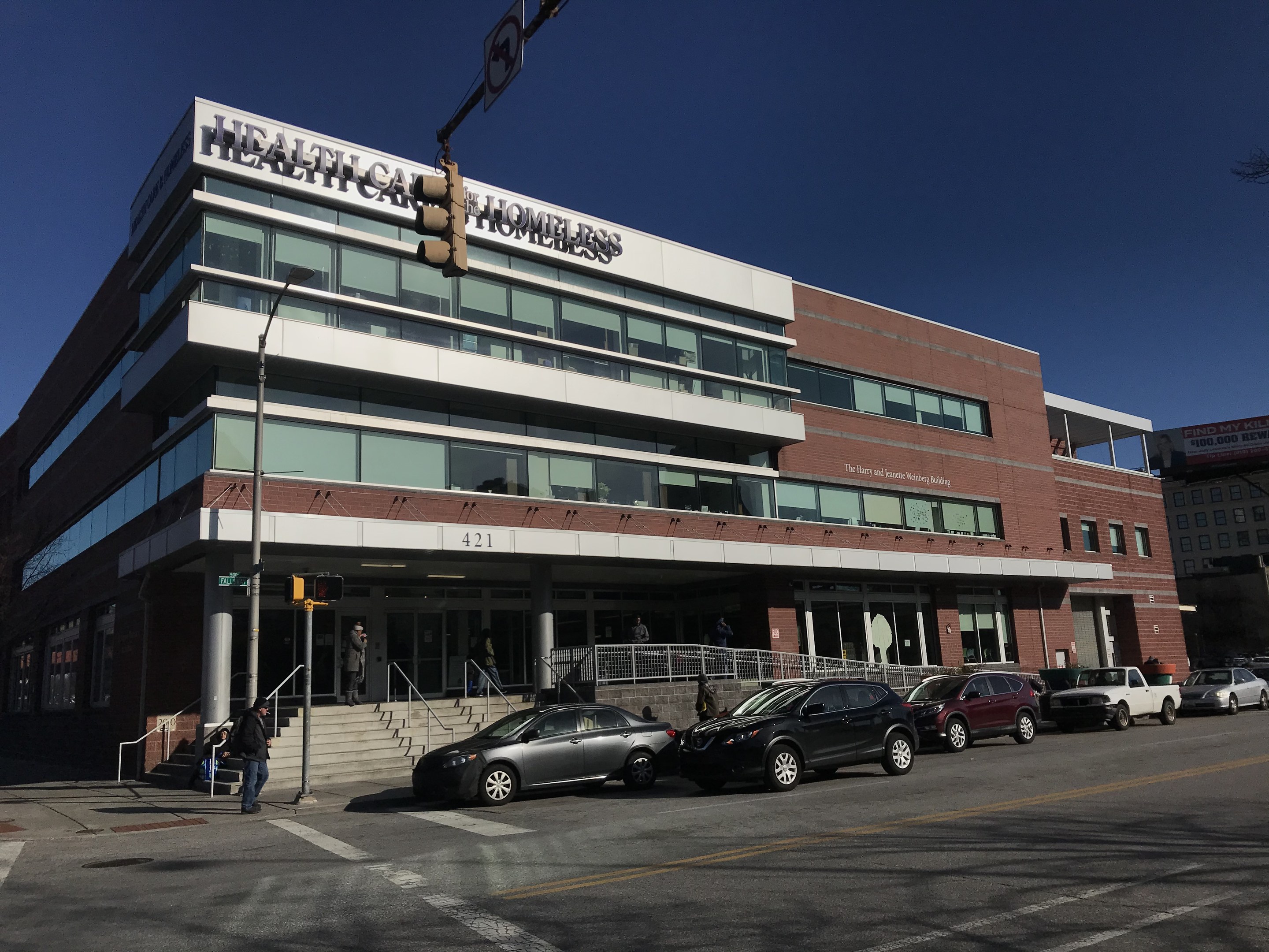 Großes Gebäude als das New York City Health Care Obdachlosenzentrum mit geparkten Autos, Fußgängern, Straßenmöblierung und Bäumen unter einem klaren Himmel.