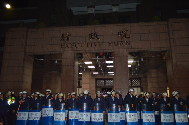 Gruppe von Polizisten in Helmen und Schilden vor einem Gebäude mit beleuchteter Wand- und Deckenbeschriftung und weiteren Gebäuden im Hintergrund.