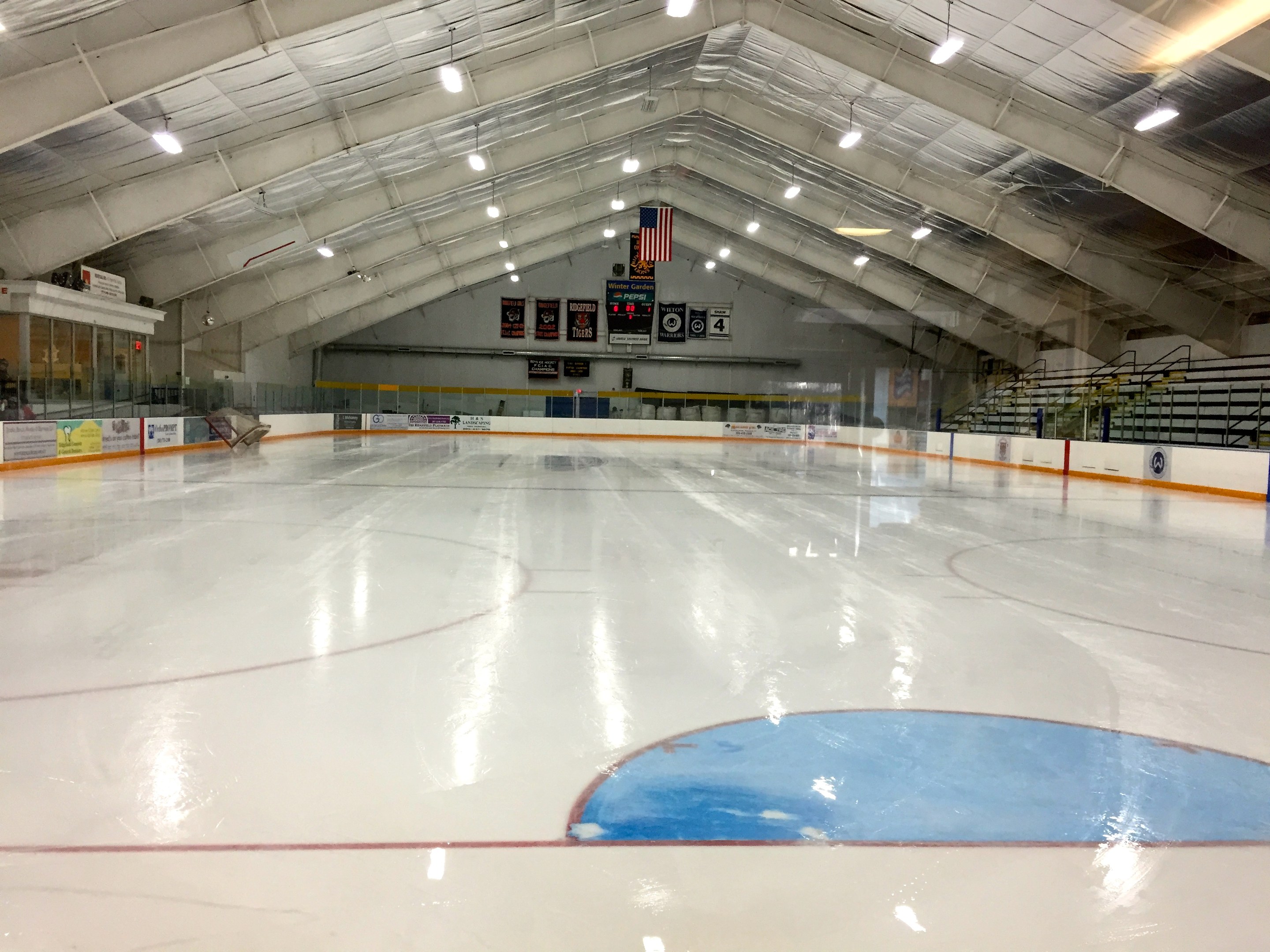 Große Indoor-Eisfläche mit Deckenbeleuchtung, Werbetafeln, einer Rückwand, Treppen mit Geländern und einigen Menschen, was auf eine Sportanlage hinweist, die likely für Hockey verwendet wird.