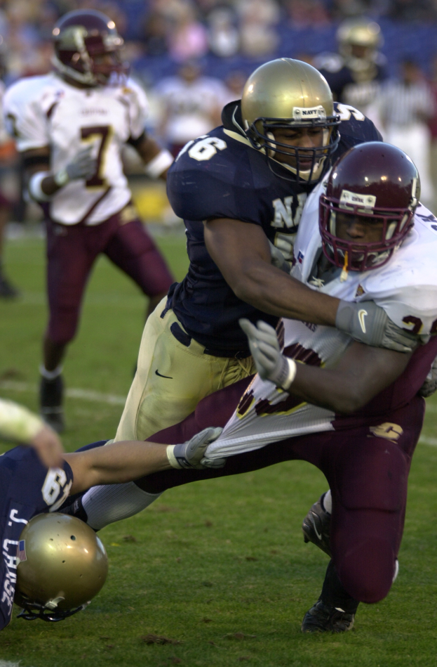 Ein Fussballspieler in einem Helm wird von einem anderen Spieler in einem Helm auf dem Feld getackelt, mit anderen Spielern und Zuschauern im Hintergrund.