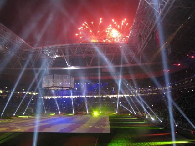 Das Olympische Stadion in London, England, nachts mit hellen Lichtern und Feuerwerken erleuchtet, voller Menschen und mit einem großen Bildschirm im Vordergrund.